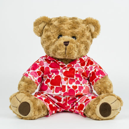 A teddy bear wearing a red and white heart-patterned pajama top and matching pajama bottoms sits on a white background.