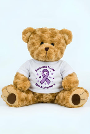 A brown teddy bear wearing a white t-shirt with a purple ribbon and the text "Someone I Love Has Alzheimer's" printed on it.