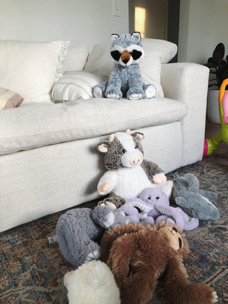 A cozy living room with a white couch and various stuffed animals, including a raccoon, cow, and bear, arranged on the floor.