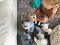 A young child is sitting on the floor surrounded by various stuffed animals, including a raccoon, a sloth, and a polar bear.