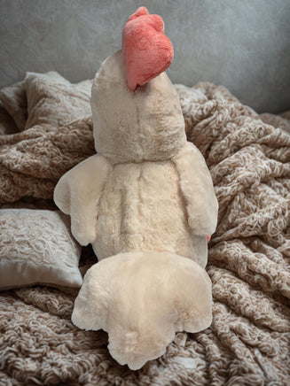 A plush toy of a chicken, with a pink beak, is sitting on a bed with a blanket.