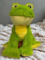 A large, plush green frog toy with yellow accents sits on a white bedspread.