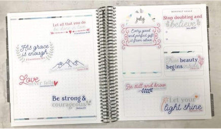 The image is an open spiral-bound notebook with various inspirational quotes and Bible verse references. The notebook is open to a page titled "July" and contains a list of Bible verse references, such as "His grace is enough" and "Be strong and courageous." The notebook also features a page titled "Monthly Goals" with a list of goals and a Bible verse reference. The overall layout and content suggest that the notebook is being used as a tool for personal