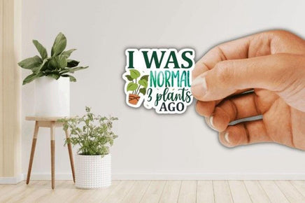 A hand holding a sticker with the text "I WAS NORMAL 3 PLANT'S AGO" in front of a wall with a potted plant and a small table.