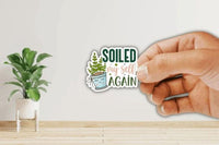 A hand holding a sticker with the text "Soiled my self again" and an illustration of a potted plant.