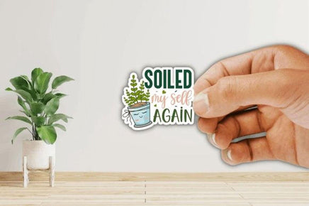 A hand holding a sticker with the text "Soiled my self again" and an illustration of a potted plant.