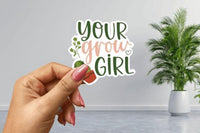 A hand holding a sticker with the text "Your grow girl" against a backdrop of a potted plant.