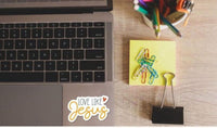 a laptop, a yellow sticky note, a binder clip, and a cup of colored pencils on a wooden surface.