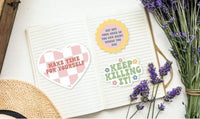 an open notebook with motivational stickers on the left page, a bouquet of purple flowers on the right side, and a straw hat on the left side.