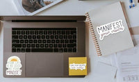 a laptop with stickers, a notebook, and some papers on a desk, along with a book and a pen.
