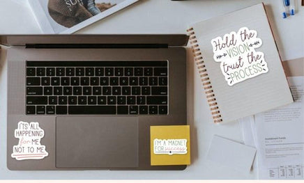 a laptop with a sticker that says "It's all happening for me" and another sticker that says "I'm a magnet for success". There is also a notebook with a sticker that says "Hold the vision trust the process".