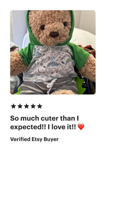 A teddy bear wearing a green hoodie and gray pants, sitting on a bed.
