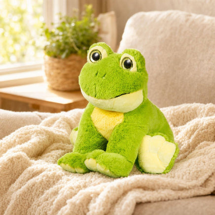 A green plush frog toy is sitting on a beige blanket on a couch, with a potted plant visible in the background.