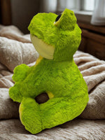 A large, plush green frog toy is sitting on a bed, with a window and beige bedding visible in the background.