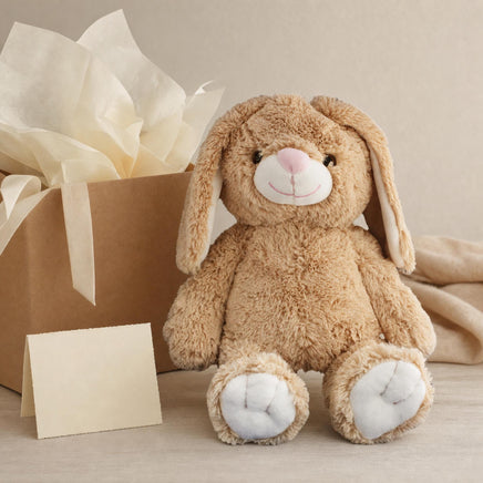 A brown stuffed bunny toy sits next to a brown gift box with a white tissue paper bow, and a blank card is placed in front of the gift box.