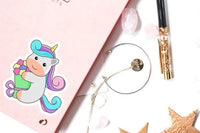 a pink notebook with a cartoon unicorn sticker, a pen, a bracelet, and some crystals on a white background.