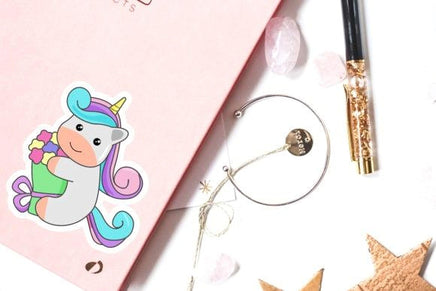 a pink notebook with a cartoon unicorn sticker, a pen, a bracelet, and some crystals on a white background.