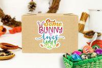 A cardboard box with a colorful sticker that says "Some Bunny loves You" is surrounded by various Easter-themed items, including a basket of colorful eggs, pine cones, and other decorations.
