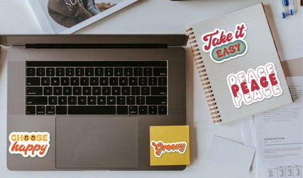 a laptop with stickers on it, along with a notebook, a pen, and some papers on a desk.