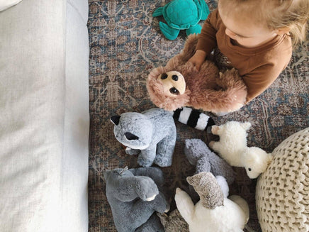A young child is sitting on the floor surrounded by various stuffed animals, including a raccoon, a sloth, and a polar bear.