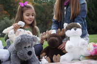 A young girl is sitting on the grass, surrounded by various stuffed animals, including a moose, a dog, and a cat.