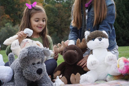 A young girl is sitting on the grass, surrounded by various stuffed animals, including a moose, a dog, and a cat.