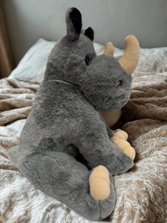 A plush toy rhinoceros with gray fur and yellow horn is sitting on a bed with a patterned blanket.