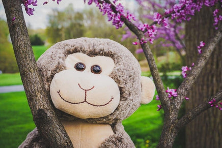 A stuffed monkey toy is sitting on a tree branch with purple flowers in the background.