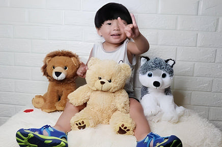 A young child sitting on a bed, surrounded by stuffed animals, including a brown lion, a yellow teddy bear, and a gray husky.