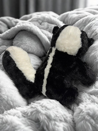 A plush toy of a skunk is sitting on a soft, fluffy surface, possibly a bed or a blanket.