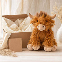 A brown, furry stuffed animal resembling a Highland cow sits next to an open cardboard box on a wooden surface, with a vase of dried flowers in the background.
