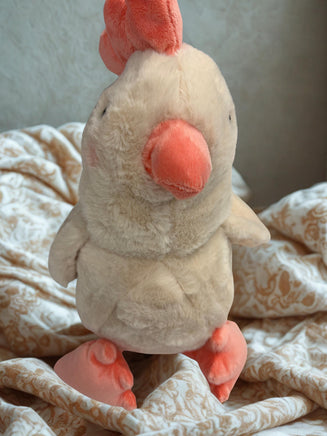A plush toy of a chicken with a pink beak and feet is sitting on a patterned fabric.