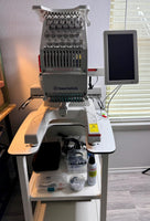 A large, white embroidery machine with various components and accessories, including a monitor and a bottle of thread, is displayed in a room with a window.