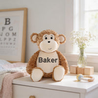 A brown stuffed monkey toy with the name "Baker" on its chest sits on a white dresser next to a vase of white flowers and a wooden block.
