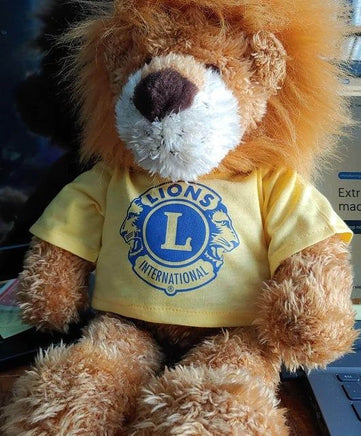 A brown teddy bear wearing a yellow t-shirt with a blue lion logo, sitting on a desk.