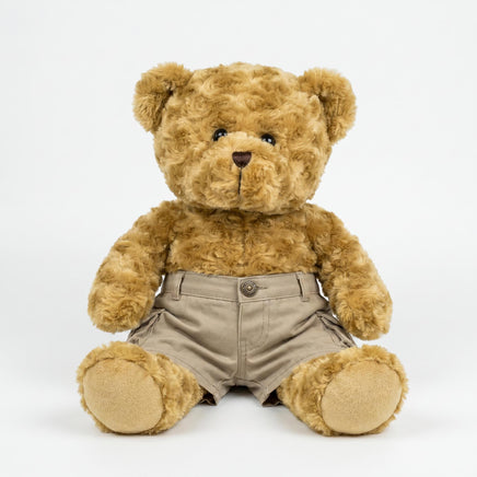 A brown teddy bear wearing khaki cargo shorts sits on a white background.