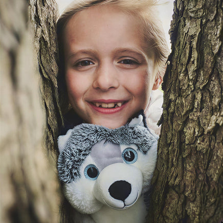 A young girl peeking out from behind a tree trunk, smiling and holding a stuffed animal.