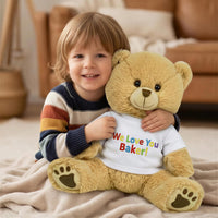 A young child is sitting on the floor, hugging a large teddy bear wearing a white shirt with the text "We Love You Baker!" printed on it.