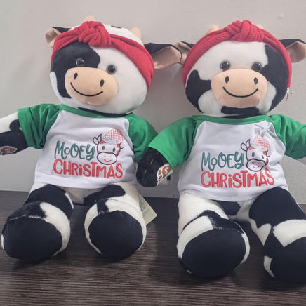 Two stuffed cows wearing Christmas-themed shirts and red bandanas are sitting next to each other.