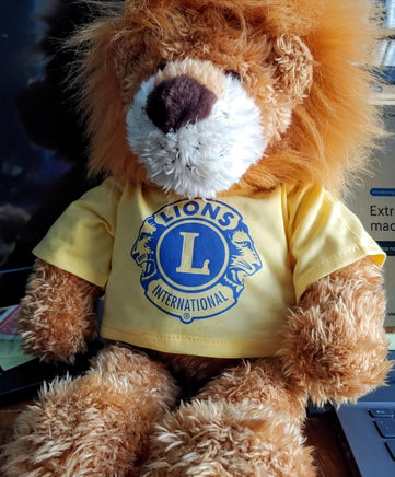 A brown teddy bear wearing a yellow t-shirt with a blue lion logo, sitting on a desk.