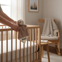 A person's hands holding a sloth plush toy with the name "Baker" on it, placed on a wooden crib.
