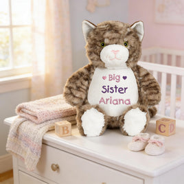 A stuffed cat toy with a personalized message sits on a white dresser in a nursery, accompanied by baby booties and a blanket.